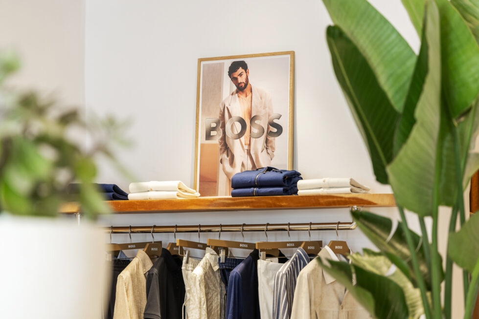 A curated selection of garments is displayed with precision, blending soft neutral tones and deep blues. The warm wood shelving and green foliage add an organic contrast, reinforcing the store’s effortlessly sophisticated and natural aesthetic.