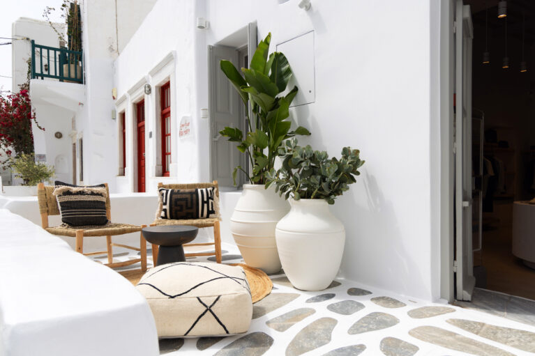 A relaxed outdoor space featuring woven seating, handcrafted ceramics, and lush greenery. The neutral palette and organic materials blend effortlessly with the traditional Cycladic architecture, creating a tranquil retreat for visitors.