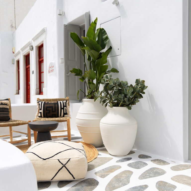 A relaxed outdoor space featuring woven seating, handcrafted ceramics, and lush greenery. The neutral palette and organic materials blend effortlessly with the traditional Cycladic architecture, creating a tranquil retreat for visitors.