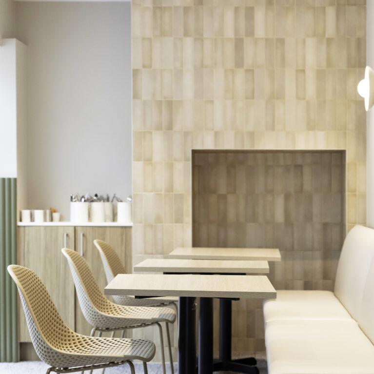 Sofa’s dining area features a neutral palette with soft beige tiles and minimalist furniture. Light wood tables and woven chairs bring warmth, while clean lines enhance the modern, inviting ambiance.