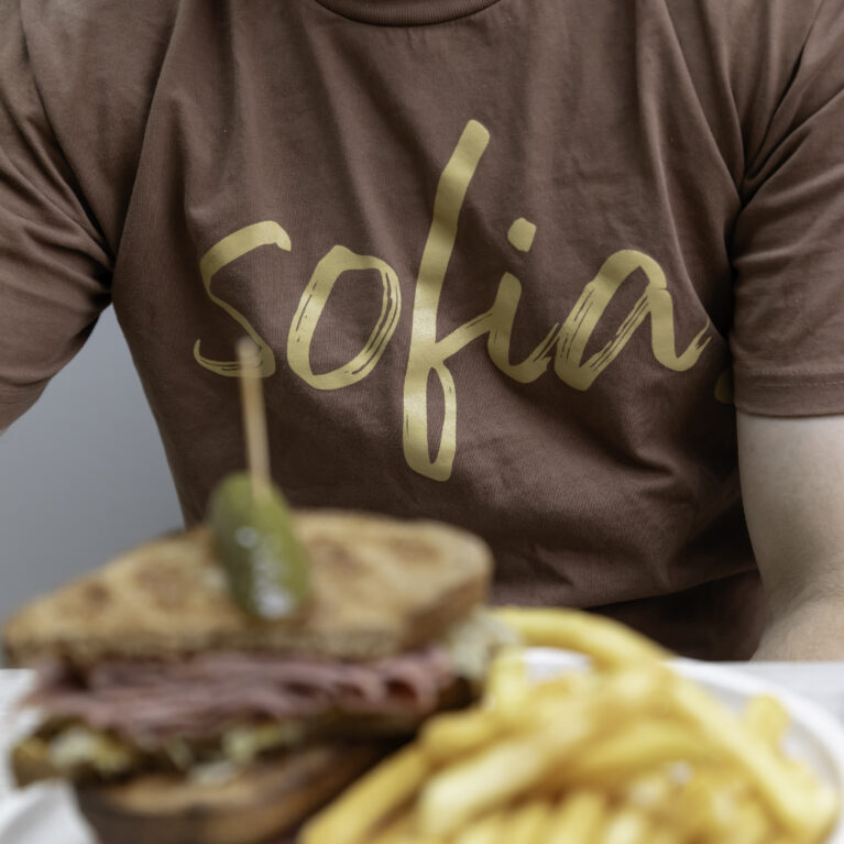 The "Sofia" logo, styled in simple handwritten typography, reflects the café's approachable branding. Set against a rich brown t-shirt and a hearty sandwich, it ties the visual identity to the warm, earthy tones of the café’s inviting atmosphere.