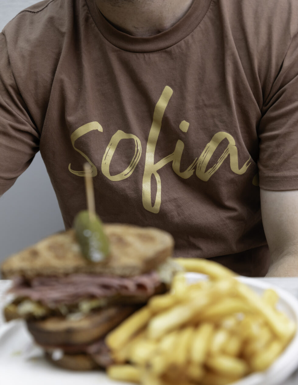 The "Sofia" logo, styled in simple handwritten typography, reflects the café's approachable branding. Set against a rich brown t-shirt and a hearty sandwich, it ties the visual identity to the warm, earthy tones of the café’s inviting atmosphere.