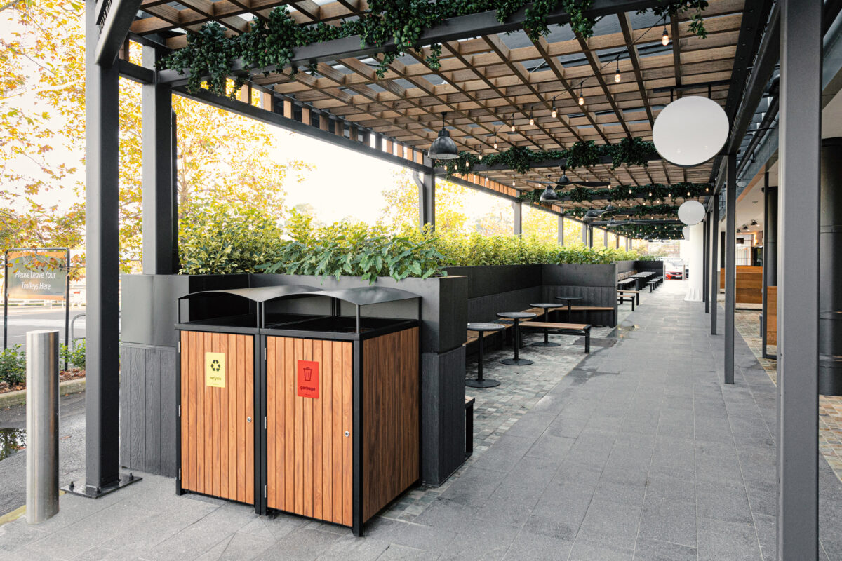 Timber clad bin enclosure, recycling station, steel arbour framework, timber lattice ceiling, outdoor dining concourse, grey stone paving, stainless steel bollard, perimeter planter boxes, festoon lighting, Sydney retail precinct.