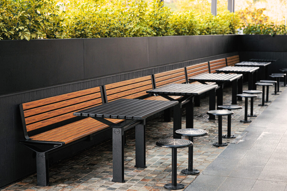 Timber banquette bench, slatted dining tables, fixed stool pedestals, cobblestone paving, perimeter hedge planter, dark wall paneling, high-density seating, outdoor dining zone, Sydney retail precinct.