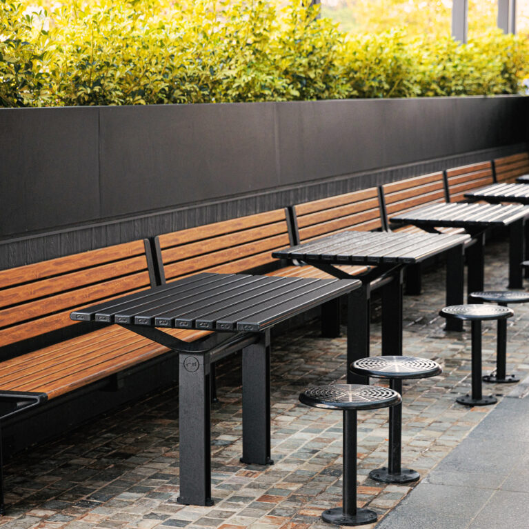 Timber banquette bench, slatted dining tables, fixed stool pedestals, cobblestone paving, perimeter hedge planter, dark wall paneling, high-density seating, outdoor dining zone, Sydney retail precinct.