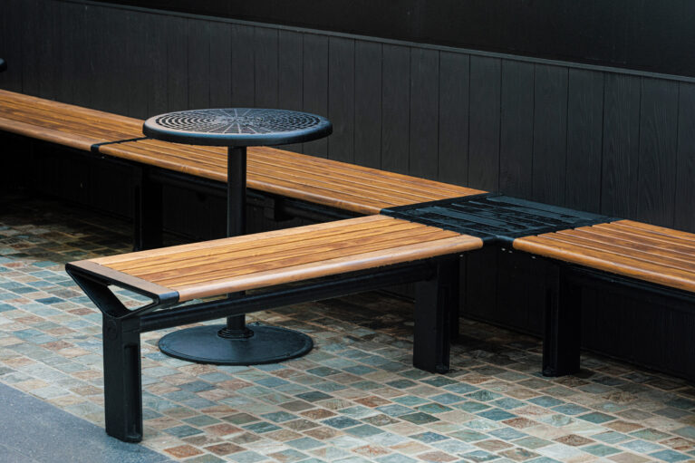 Modular timber bench, L-shape seating layout, cast iron pedestal table, perforated metal top, dark wall cladding, multi-tonal cobble pavers, urban furniture design, precinct alfresco zone, Sydney retail design.