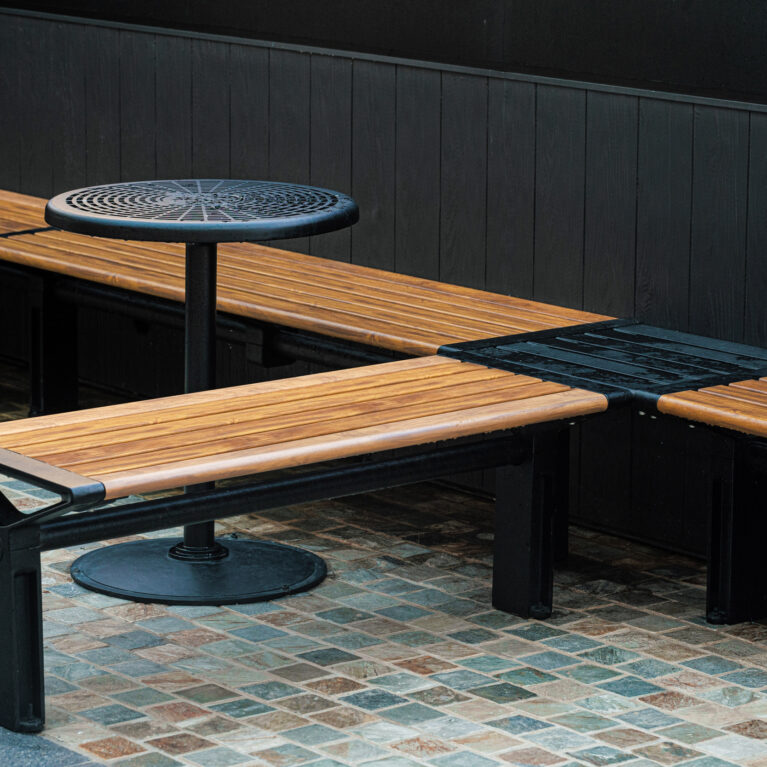 Modular timber bench, L-shape seating layout, cast iron pedestal table, perforated metal top, dark wall cladding, multi-tonal cobble pavers, urban furniture design, precinct alfresco zone, Sydney retail design.