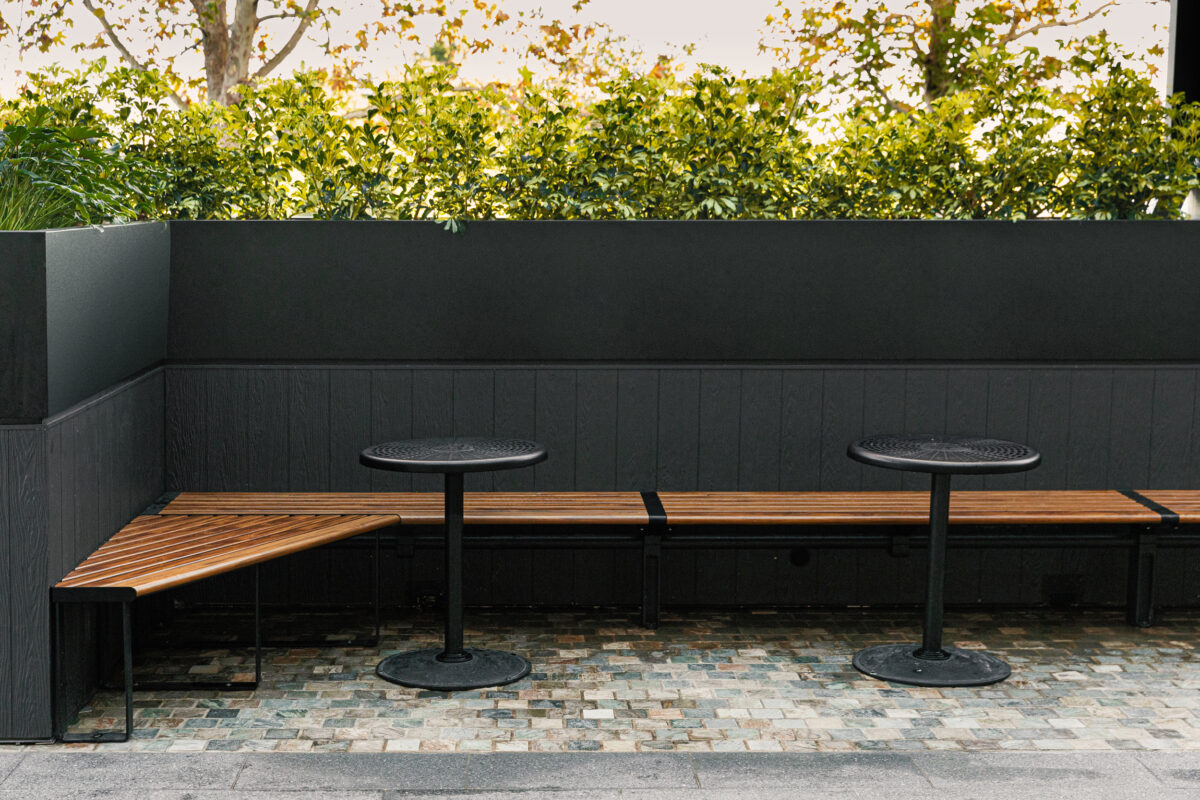 Timber banquette seating, angled bench corner, built-in planter box, dark exterior wainscoting, black pedestal tables, cobble paving, perimeter hedging, alfresco nook, Sydney retail design.
