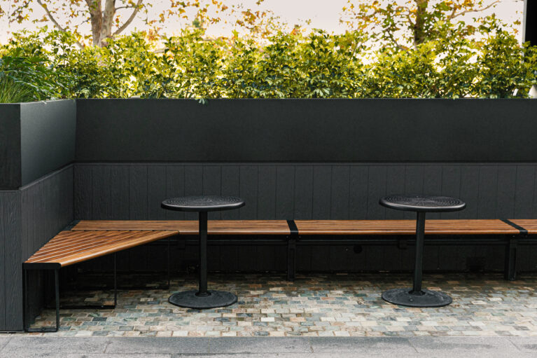Timber banquette seating, angled bench corner, built-in planter box, dark exterior wainscoting, black pedestal tables, cobble paving, perimeter hedging, alfresco nook, Sydney retail design.