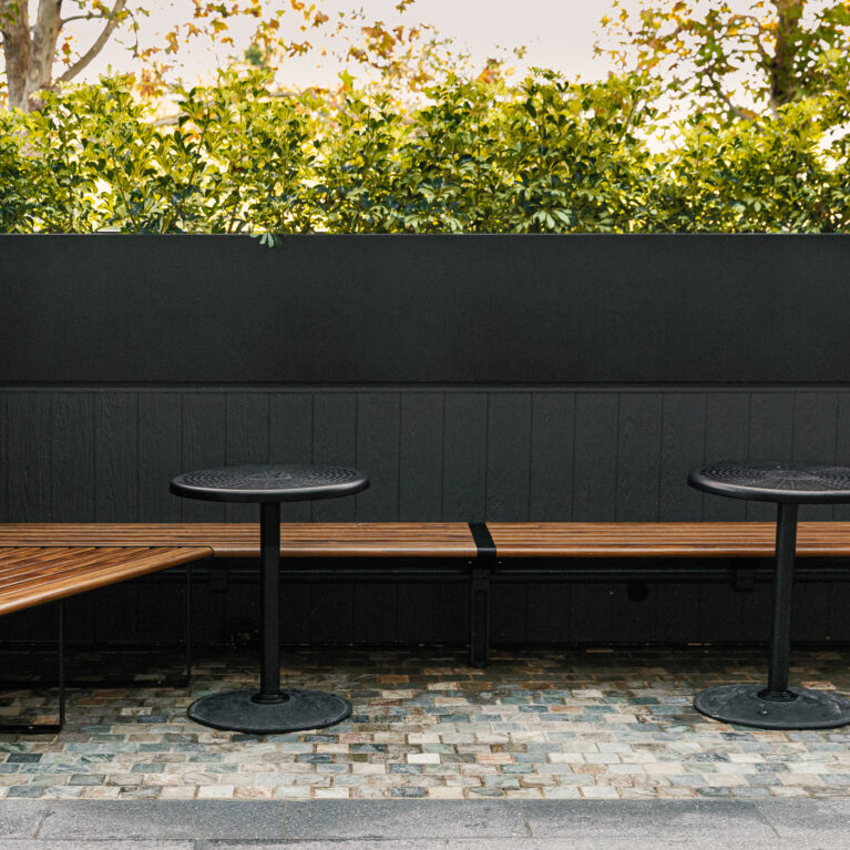 Timber banquette seating, angled bench corner, built-in planter box, dark exterior wainscoting, black pedestal tables, cobble paving, perimeter hedging, alfresco nook, Sydney retail design.