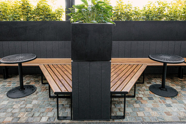 Symmetrical seating layout, central planter plinth, angled timber benches, cast iron pedestal tables, dark wainscoting, cobblestone flooring, biophilic spatial anchor, precinct outdoor dining, Sydney retail.