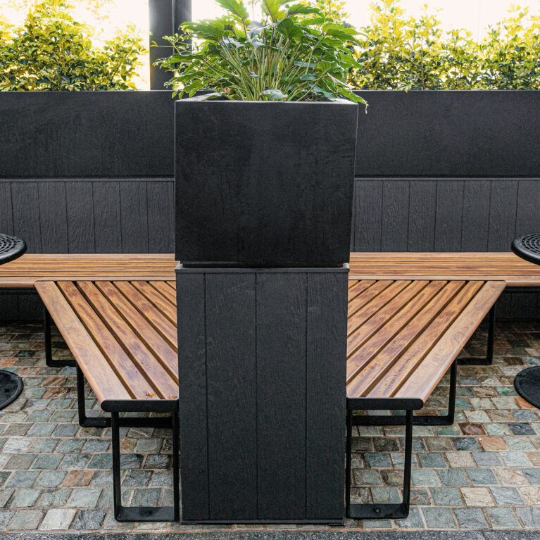 Symmetrical seating layout, central planter plinth, angled timber benches, cast iron pedestal tables, dark wainscoting, cobblestone flooring, biophilic spatial anchor, precinct outdoor dining, Sydney retail.