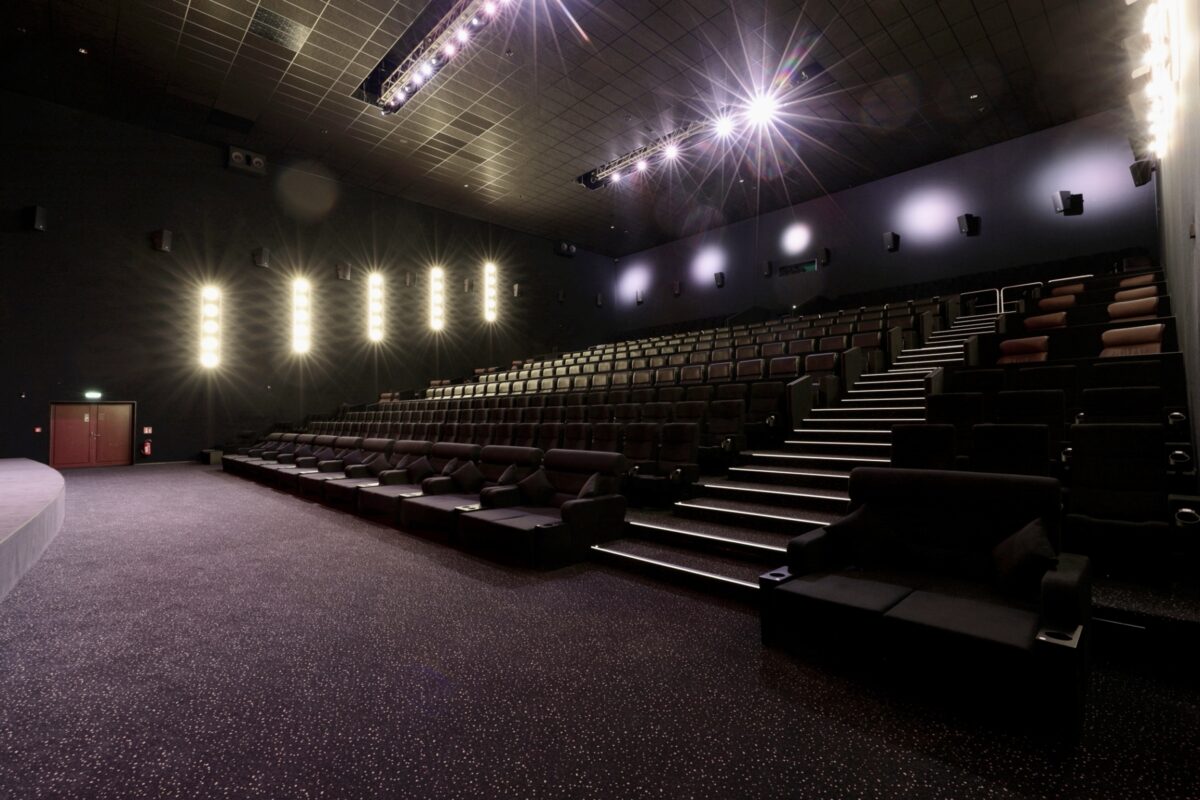 Cinema stadium seating, acoustic wall panels, stepped theater layout, premium plush armchairs, vertical led wall sconces, dark commercial carpeting, architectural overhead spotlighting, performance venue interior.