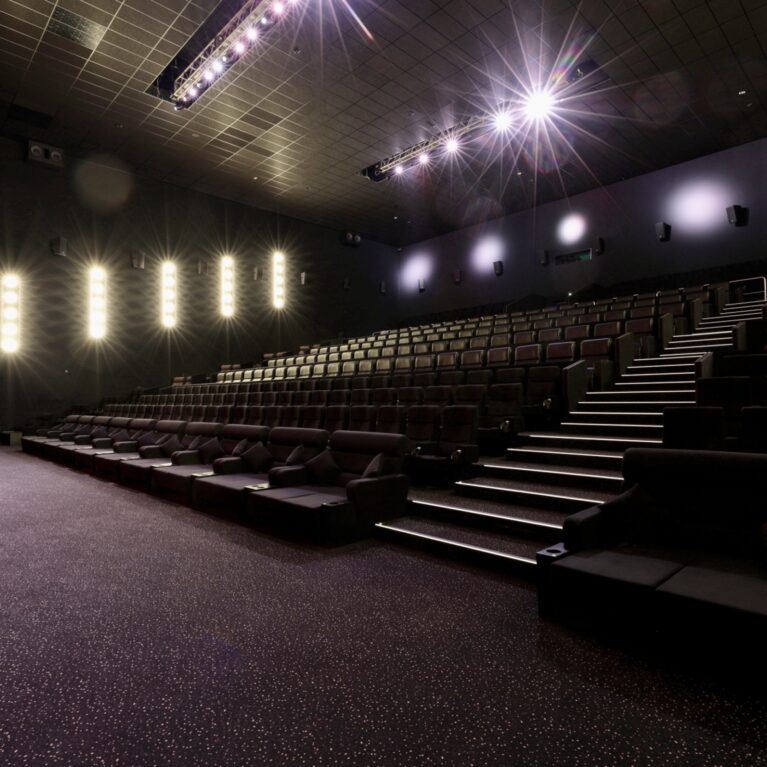 Cinema stadium seating, acoustic wall panels, stepped theater layout, premium plush armchairs, vertical led wall sconces, dark commercial carpeting, architectural overhead spotlighting, performance venue interior.