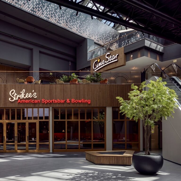 Indoor plaza layout, architectural planter seating, commercial timber framing, multi-level facade alignment, illuminated brand signage, dark steel roof truss, large format floor tiling, landscape interior accents.