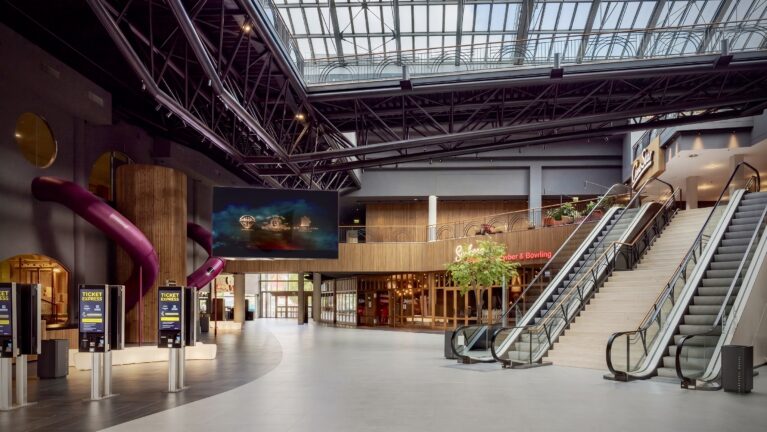 Cinema entry foyer, dual commercial escalators, industrial steel roof truss, architectural timber cladding, self-service ticketing kiosks, large format digital signage, indoor playground slides, open plan transit layout.