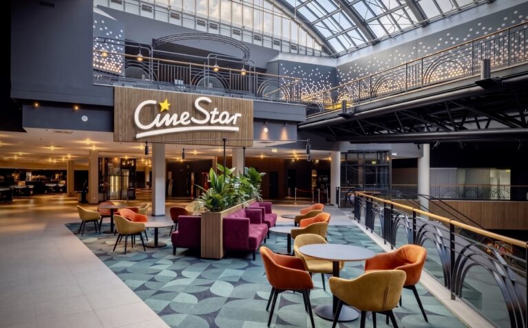 Cinema atrium interior, commercial lounge seating, modern theater mezzanine, open plan lobby design, pattern carpet tiles, upholstered armchairs, timber cladding branding, glass atrium roof, architectural balustrade.