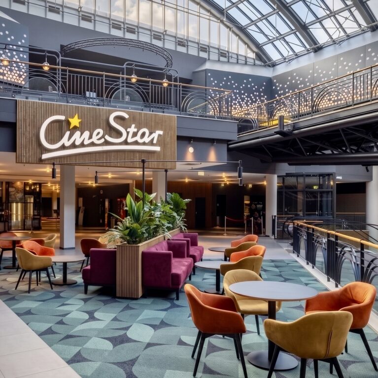 Cinema atrium interior, commercial lounge seating, modern theater mezzanine, open plan lobby design, pattern carpet tiles, upholstered armchairs, timber cladding branding, glass atrium roof, architectural balustrade.