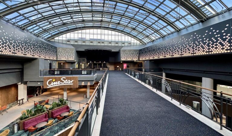 Architectural sky bridge, glass barrel vault roof, suspended pedestrian walkway, dark acoustic carpeting, decorative perforated metal, structural steel trussing, mezzanine interior overlook, cinema atrium design.
