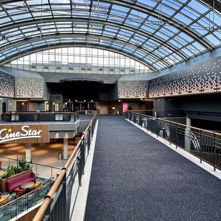 Architectural sky bridge, glass barrel vault roof, suspended pedestrian walkway, dark acoustic carpeting, decorative perforated metal, structural steel trussing, mezzanine interior overlook, cinema atrium design.