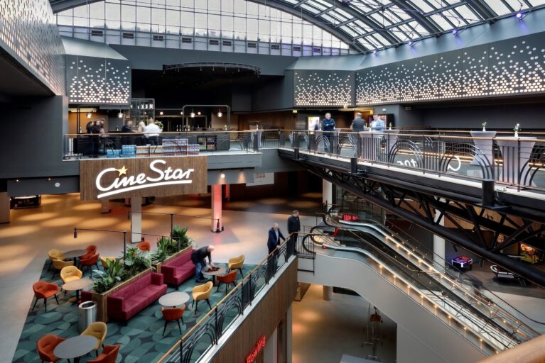 Multi-story cinema atrium, architectural glass vaulted roof, indoor promenade mezzanine, structural steel trusses, tiered bar level, custom glass balustrades, integrated escalator shafts, perforated wall paneling.