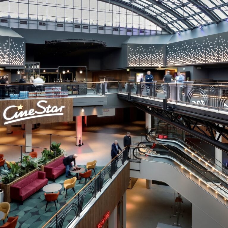 Multi-story cinema atrium, architectural glass vaulted roof, indoor promenade mezzanine, structural steel trusses, tiered bar level, custom glass balustrades, integrated escalator shafts, perforated wall paneling.