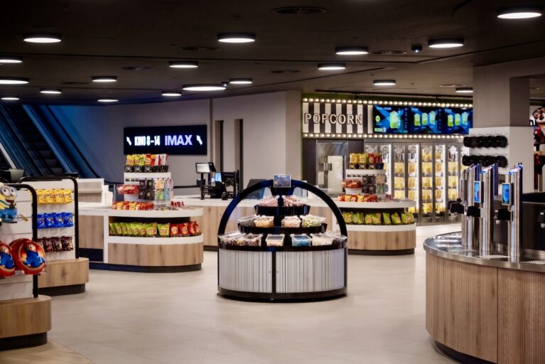 Cinema marketplace layout, retail display islands, concession stand design, timber clad counters, grab and go snack station, commercial pendant lighting, digital signage, theater concourse interior, terrazzo style flooring, modern box office retail.