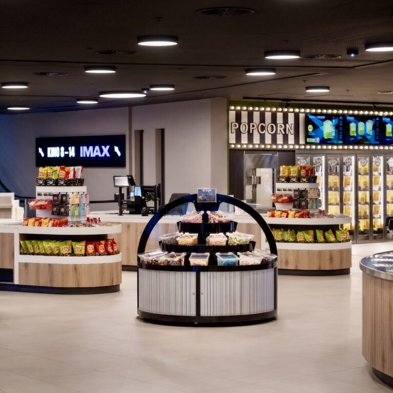 Cinema marketplace layout, retail display islands, concession stand design, timber clad counters, grab and go snack station, commercial pendant lighting, digital signage, theater concourse interior, terrazzo style flooring, modern box office retail.
