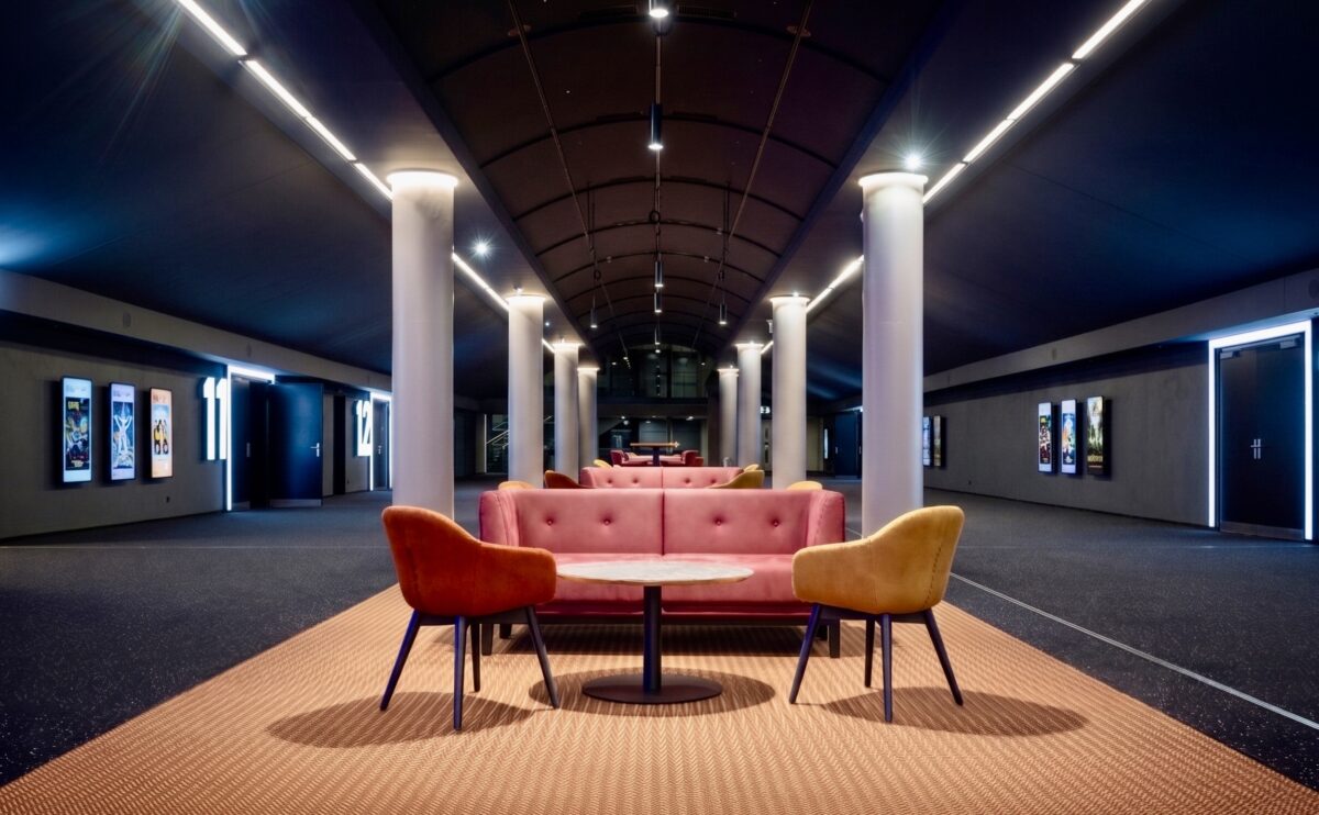 Modern cinema lobby design, hospitality lounge seating, barrel vault ceiling, linear track lighting, commercial carpet flooring, mid-century modern armchairs, tufted plush sofa, architectural columns, cinematic corridor lighting, theater entrance signage.
