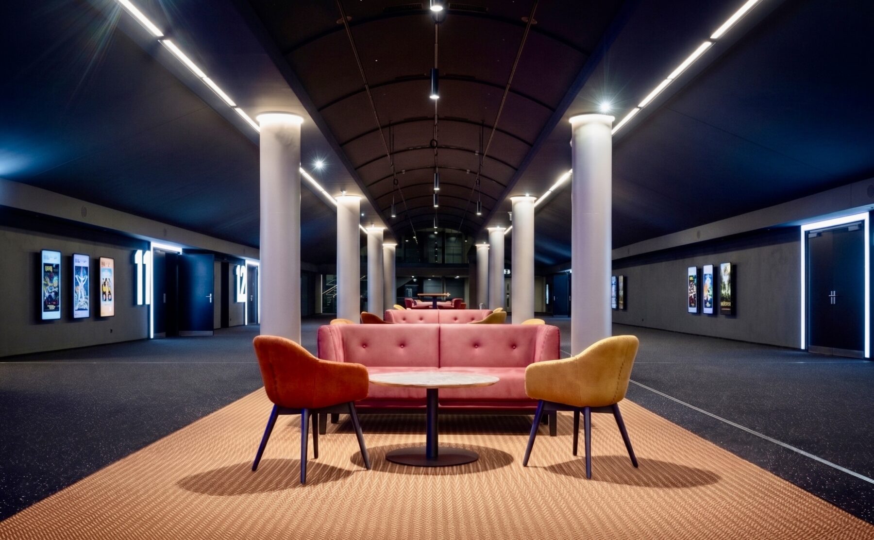 Modern cinema lobby design, hospitality lounge seating, barrel vault ceiling, linear track lighting, commercial carpet flooring, mid-century modern armchairs, tufted plush sofa, architectural columns, cinematic corridor lighting, theater entrance signage.