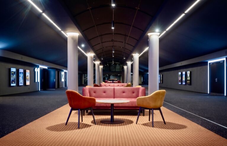Modern cinema lobby design, hospitality lounge seating, barrel vault ceiling, linear track lighting, commercial carpet flooring, mid-century modern armchairs, tufted plush sofa, architectural columns, cinematic corridor lighting, theater entrance signage.