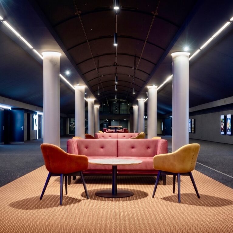 Modern cinema lobby design, hospitality lounge seating, barrel vault ceiling, linear track lighting, commercial carpet flooring, mid-century modern armchairs, tufted plush sofa, architectural columns, cinematic corridor lighting, theater entrance signage.