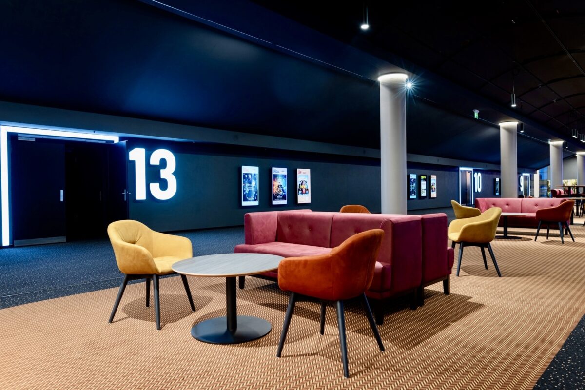 Cinema corridor seating, theater portal signage, illuminated room numbers, commercial hallway lounge, velvet club chairs, modular waiting sofa, textured carpet runners, structural pillars, architectural lighting design, hospitality lounge furniture.