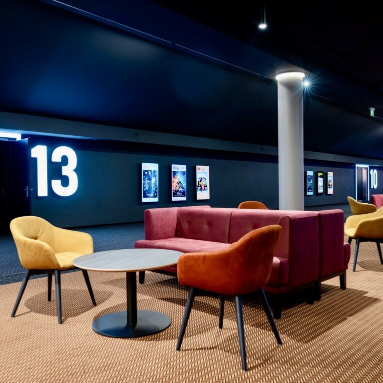 Cinema corridor seating, theater portal signage, illuminated room numbers, commercial hallway lounge, velvet club chairs, modular waiting sofa, textured carpet runners, structural pillars, architectural lighting design, hospitality lounge furniture.