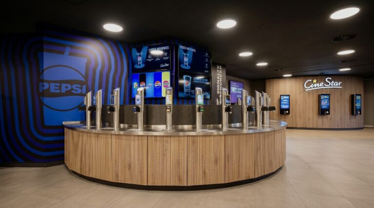 Cinema concession stand, self-service drink station, curved wooden counter, stainless steel beverage dispensers, digital menu boards, recessed ceiling downlights, oak wall paneling, ticketing kiosks, open plan marketplace layout, commercial hospitality design.
