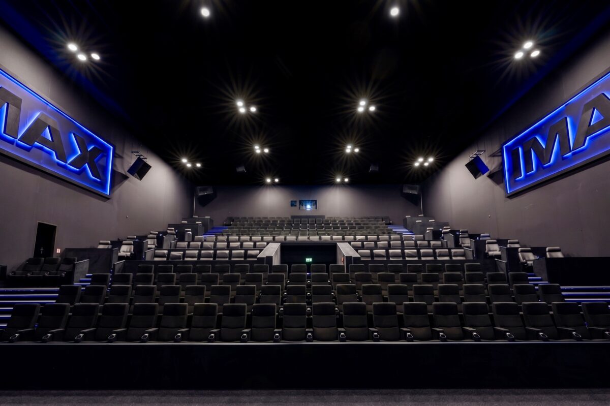 IMAX cinema auditorium, tiered stadium seating, commercial theater chairs, acoustic wall panels, blue neon signage, recess ceiling spotlights, architectural sound system, modern cinema interior, dark acoustic finishes, premium viewing hall.