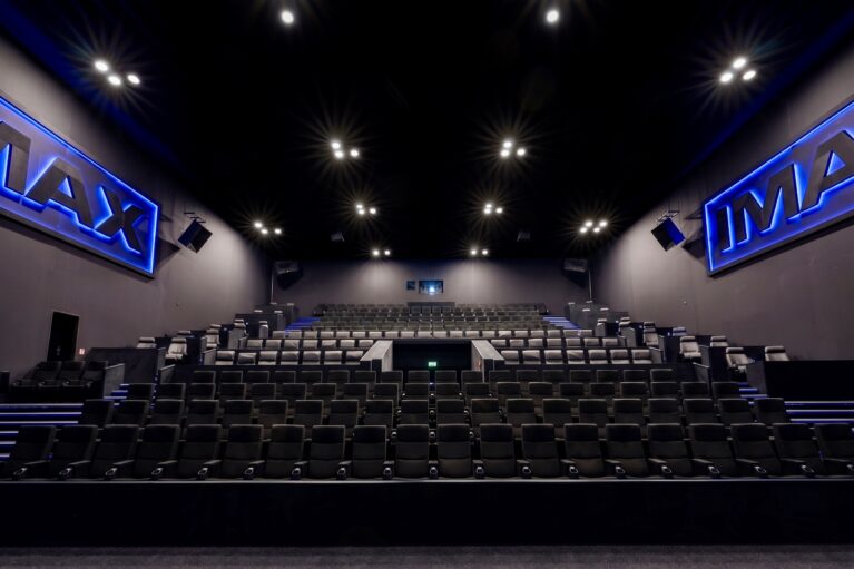 IMAX cinema auditorium, tiered stadium seating, commercial theater chairs, acoustic wall panels, blue neon signage, recess ceiling spotlights, architectural sound system, modern cinema interior, dark acoustic finishes, premium viewing hall.