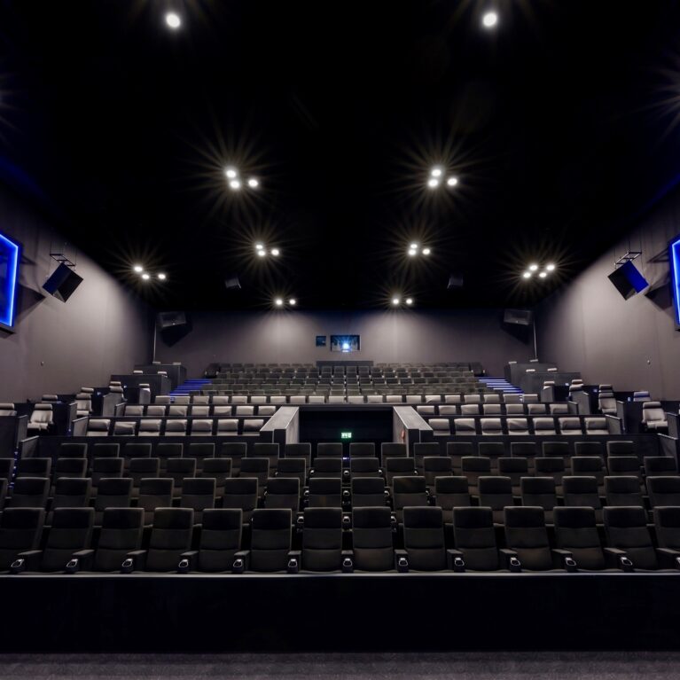 IMAX cinema auditorium, tiered stadium seating, commercial theater chairs, acoustic wall panels, blue neon signage, recess ceiling spotlights, architectural sound system, modern cinema interior, dark acoustic finishes, premium viewing hall.