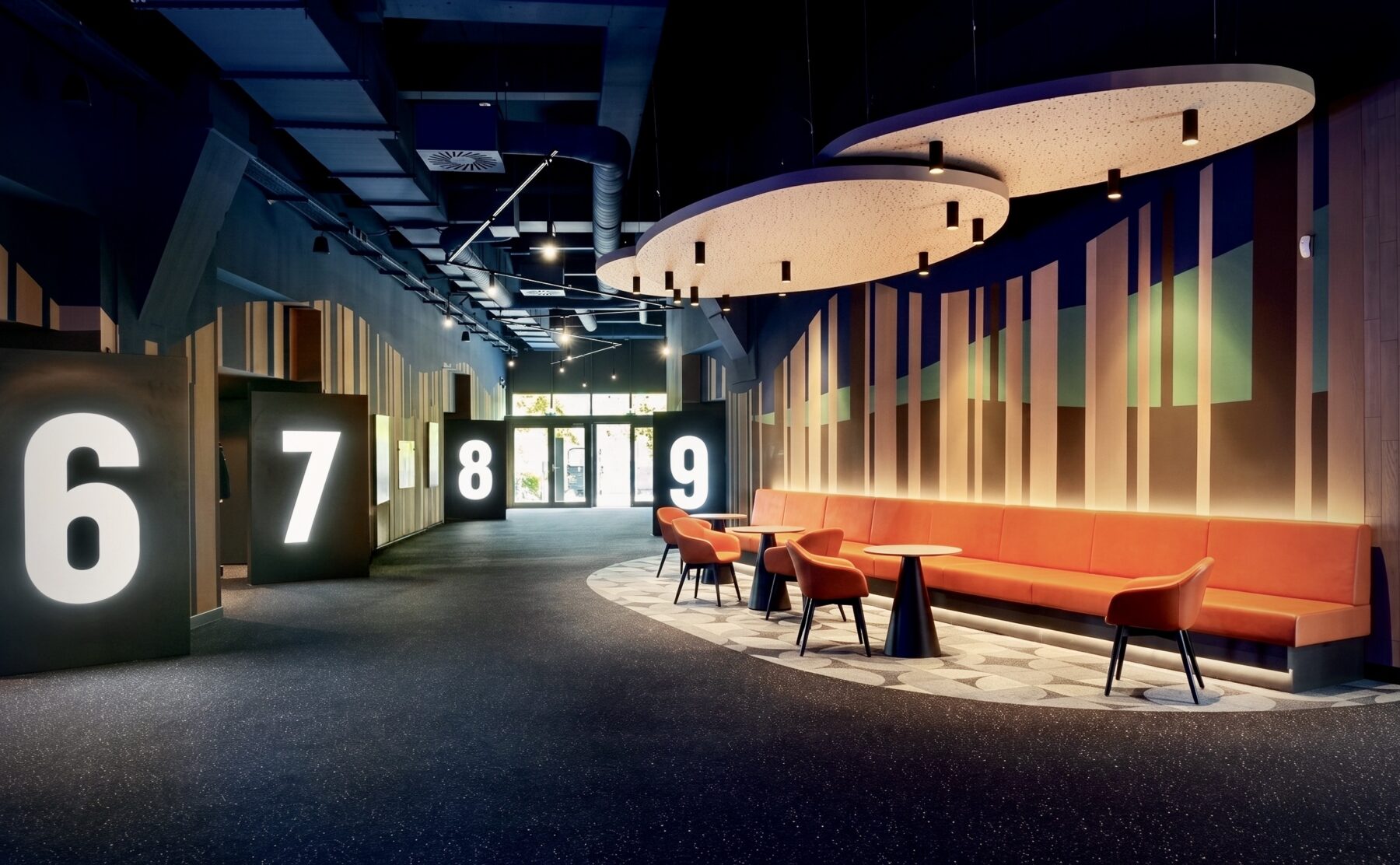 Illuminated monolithic wayfinding, acoustic floating ceiling panels, integrated banquette bench, dark speckled carpeting, vertical timber slat wall, indirect LED cove lighting, low-glare recessed downlights, commercial cinema concourse layout, customized floor pattern inlay.