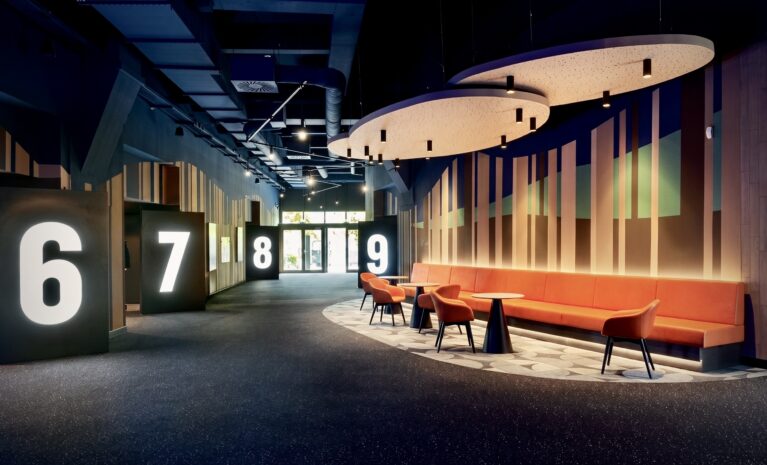 Illuminated monolithic wayfinding, acoustic floating ceiling panels, integrated banquette bench, dark speckled carpeting, vertical timber slat wall, indirect LED cove lighting, low-glare recessed downlights, commercial cinema concourse layout, customized floor pattern inlay.