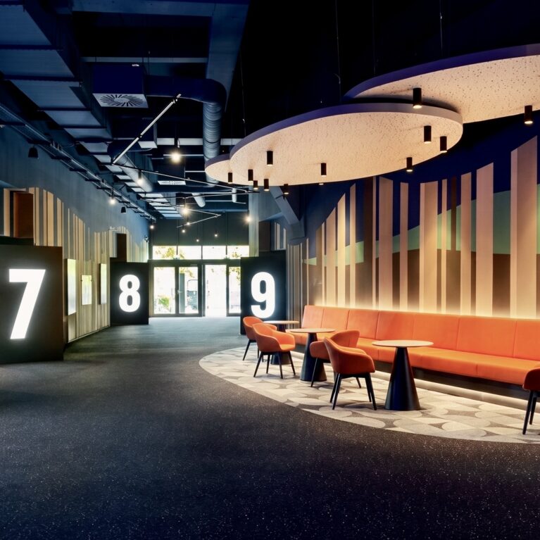 Illuminated monolithic wayfinding, acoustic floating ceiling panels, integrated banquette bench, dark speckled carpeting, vertical timber slat wall, indirect LED cove lighting, low-glare recessed downlights, commercial cinema concourse layout, customized floor pattern inlay.