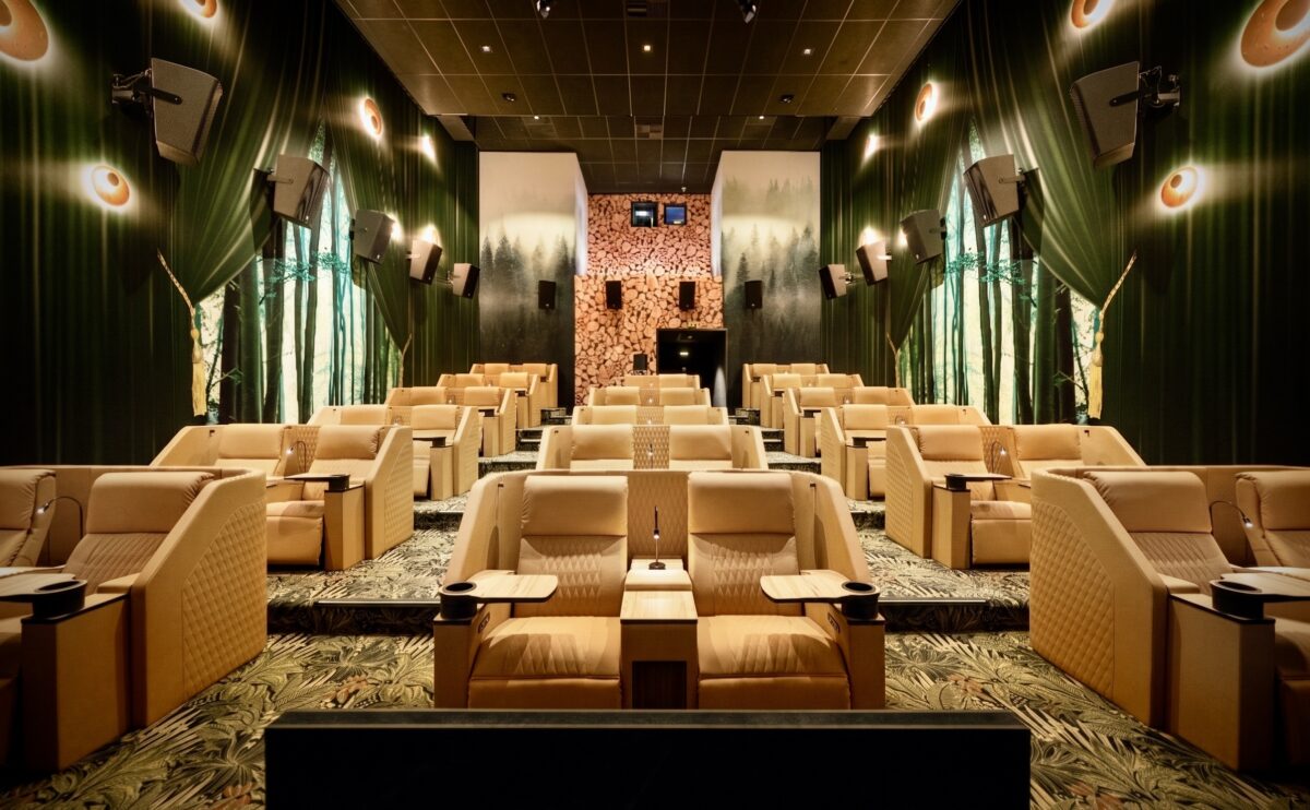 Beige quilted leather recliners, botanical patterned carpet, forest wall mural, timber log accent wall, directional seat lamps, surround sound speakers, tiered boutique auditorium, warm ambient sconces