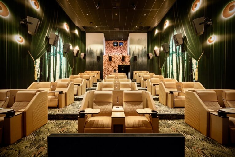 Beige quilted leather recliners, botanical patterned carpet, forest wall mural, timber log accent wall, directional seat lamps, surround sound speakers, tiered boutique auditorium, warm ambient sconces
