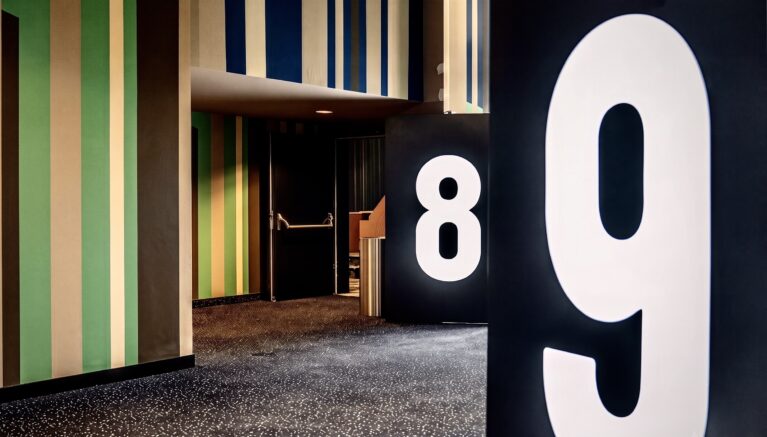 Oversized illuminated typography, black wayfinding pylons, vertical striped wall cladding, dark speckled carpet, acoustic light lock portal, recessed door alcove, graphic corridor threshold