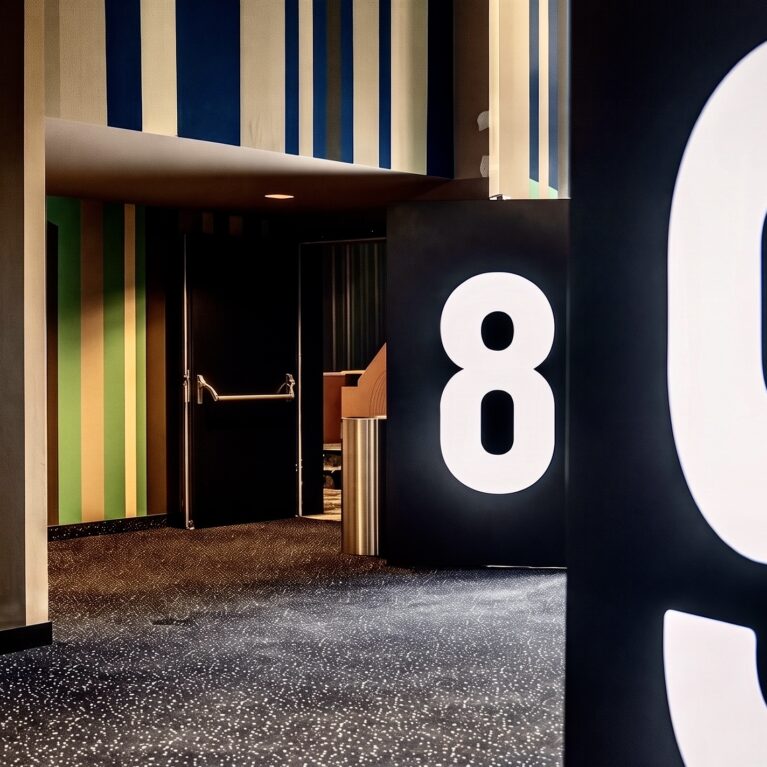 Oversized illuminated typography, black wayfinding pylons, vertical striped wall cladding, dark speckled carpet, acoustic light lock portal, recessed door alcove, graphic corridor threshold