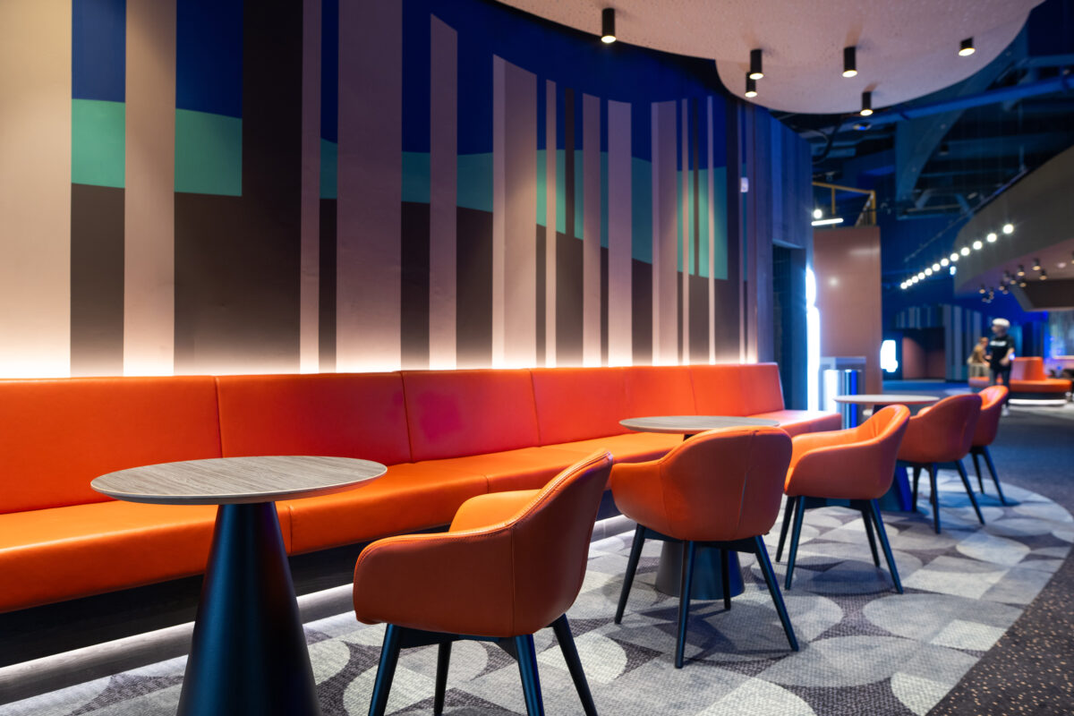 Orange leather banquette, low-slung tub chairs, conical pedestal tables, hidden cove lighting, geometric wall graphic, acoustic ceiling panel, patterned carpet inlay, cinema waiting lounge