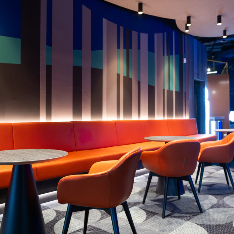 Orange leather banquette, low-slung tub chairs, conical pedestal tables, hidden cove lighting, geometric wall graphic, acoustic ceiling panel, patterned carpet inlay, cinema waiting lounge