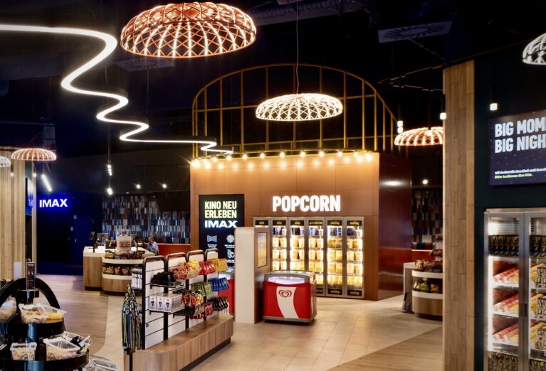 Popcorn warming cabinets, copper feature wall, festoon bulb lighting, open retail shelving, woven cage pendants, continuous LED ribbon, tiled concession floor, IMAX entry portal, commercial retail design