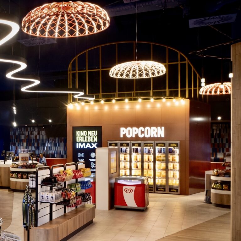 Popcorn warming cabinets, copper feature wall, festoon bulb lighting, open retail shelving, woven cage pendants, continuous LED ribbon, tiled concession floor, IMAX entry portal, commercial retail design