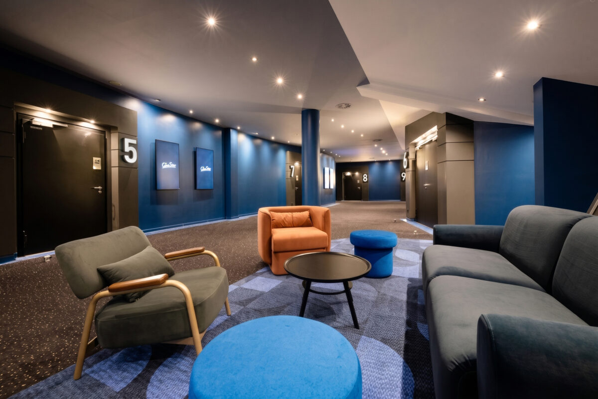 Cinema concourse lounge, breakout seating cluster, velvet upholstery, patterned modular carpet, recessed downlights, deep blue wall paint, wayfinding signage, auditorium entrance portal, commercial entertainment interior